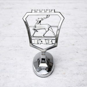May include: A chrome-finished GAZ emblem featuring a stylized deer within a shield-shaped frame. The emblem is mounted on a rounded base. The word "ГАЗ" is displayed in capital letters below the deer.