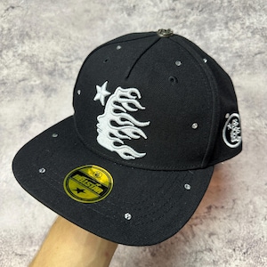 May include: Black baseball cap with a flat brim, featuring a white flame design and star. The cap has a small metal emblem on top and is embellished with small, clear rhinestones. A gold and yellow sticker with the text "HELLSTAR" is on the brim.