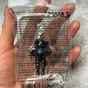 May include: A black KAWS Companion figure in a clear plastic package. The figure has a stylized, blocky design with rounded features. The packaging includes the KAWS logo and the text "KAWS COMPANION".