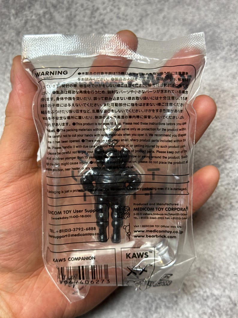 May include: A black KAWS Companion figure in a clear plastic package. The figure has a stylized, blocky design with rounded features. The packaging includes the KAWS logo and the text "KAWS COMPANION".
