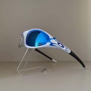 May include: White and blue sunglasses with blue mirrored lenses. The sunglasses feature a blue flame design on the frame and black arms. The sunglasses are displayed on a clear plastic stand.
