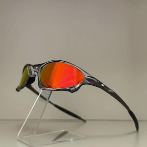 Peut inclure: Des lunettes de soleil grises avec des verres rouges vifs, présentées sur un présentoir en acrylique transparent. Les lunettes de soleil ont un design sportif, avec des branches incurvées et une monture profilée. Les verres reflètent une couleur orange vif.
