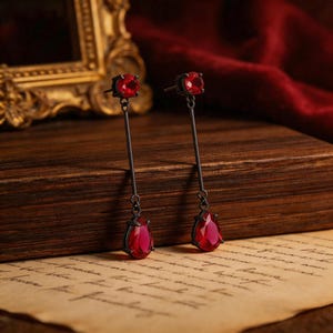May include: A pair of drop earrings featuring a dark metal post with a round red faceted stone at the top and a larger pear-shaped red faceted stone dangling from a thin vertical bar. The earrings have a dark finish and a classic drop design.