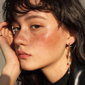 May include: A gold-toned drop earring featuring a crescent moon stud at the top, followed by a rectangular clear crystal, a square-cut clear crystal, and a small star-shaped charm at the bottom, all connected by a thin gold-toned metal chain.