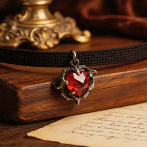 May include: A black woven choker necklace featuring a central pendant with a faceted red heart-shaped stone encased in an antique brass-toned metal frame that resembles twisted branches or thorns, suspended from a small metal jump ring.