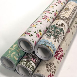May include: Five rolls of fabric featuring various patterns including pink and yellow floral prints, a green leaf design, a butterfly motif, and a botanical illustration with dark red and brown accents on a light beige textured background.