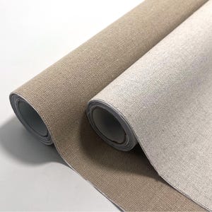 Puede incluir: Dos rollos de tela con una textura tejida visible. Un rollo es de color marrón claro medio, mientras que el otro rollo es de un color blanco roto o crema más claro. Ambos rollos son cilíndricos y muestran la estructura del material textil.
