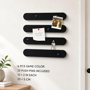 May include: Four black felt wall-mounted display strips with rounded ends. Each strip measures 33 by 5 cm. The set includes 25 push pins in various colours. The strips are used to hold a photograph, a note with handwritten text, and a set of keys.