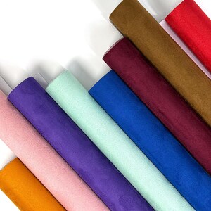 May include: A collection of eight fabric rolls arranged diagonally, featuring a variety of colors including orange, light pink, purple, mint green, royal blue, deep burgundy, brown, and bright red, all with a soft, matte, suede-like texture.