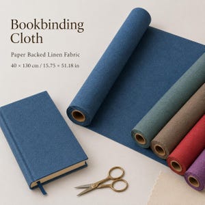 Könnte beinhalten: Buchbinderleinen, papierkaschiertes Leinen mit den Maßen 40 x 130 cm, dargestellt als blaue Rolle und gebundenes blaues Buch, neben Rollen in Grün, Braun, Rot und Violett sowie einer Schere aus goldfarbenem Metall auf der Oberfläche.