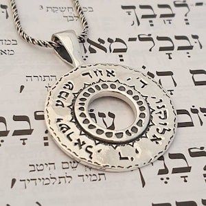 May include: A circular silver pendant on a twisted silver chain. The pendant features a central open circle surrounded by a textured border and an outer ring engraved with Hebrew text. The metal has a polished finish with darkened recessed areas.