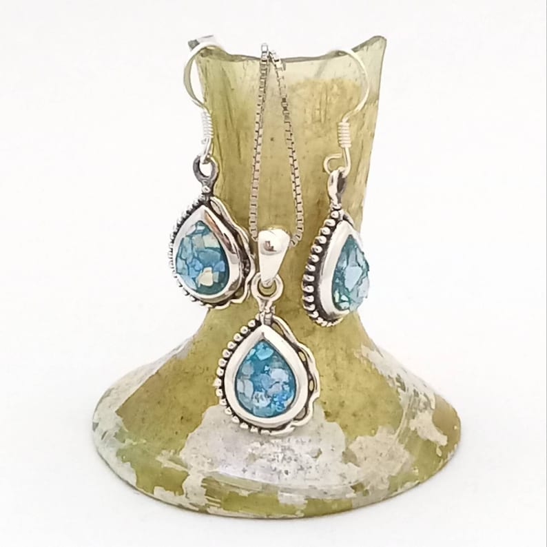 May include: A silver jewelry set featuring a teardrop pendant necklace and matching earrings. Each piece has a silver frame with a beaded edge, surrounding a light blue, mosaic-like stone. The set is displayed on a light green, textured vase.