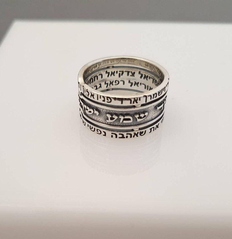 May include: A silver ring with Hebrew text inscribed around the band. The text is in a traditional Hebrew font and is arranged in four lines.