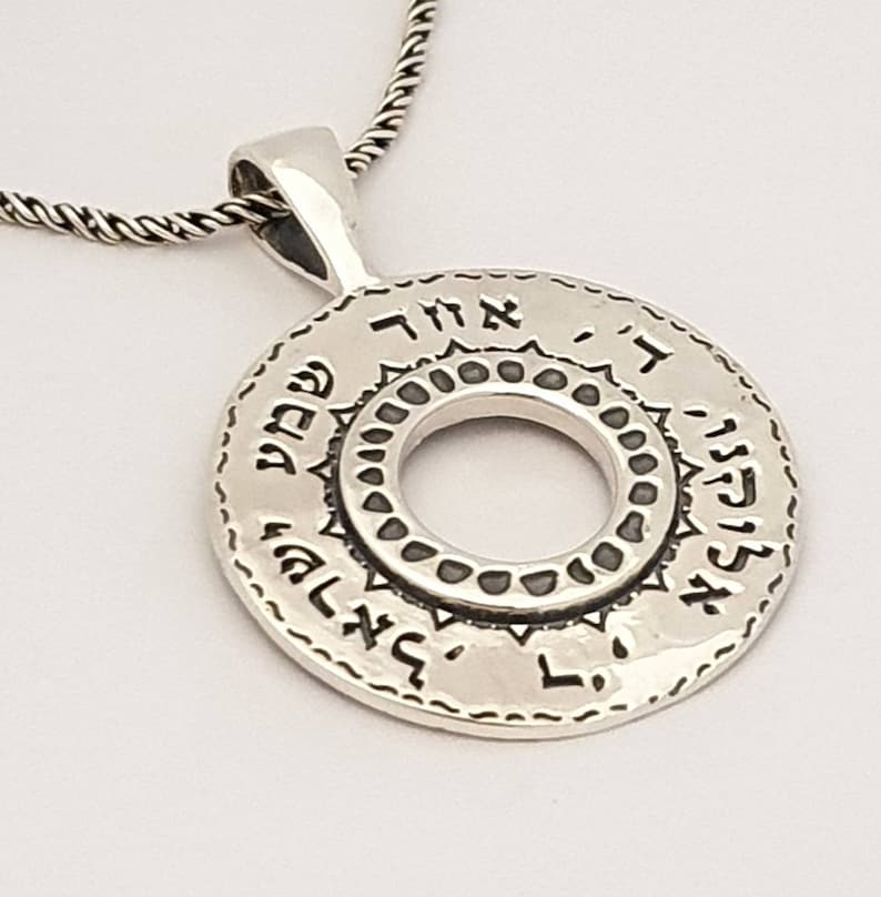 May include: A circular silver pendant featuring Hebrew text engraved along the outer rim and a central cutout surrounded by a decorative border. The pendant hangs from a twisted silver chain, showcasing a polished metallic finish throughout.