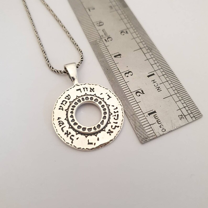 May include: A silver circular pendant necklace featuring Hebrew text engraved along the outer rim and a decorative inner ring pattern. A metal ruler placed next to the pendant shows the circular piece has a diameter of approximately 1 inch.
