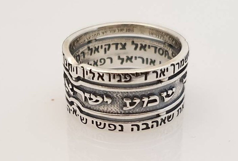 May include: A silver ring with Hebrew text inscribed around the band. The text reads "מיאל עזריאל תמניאל", "חסדיאל צדקיאלרי", "שמרך יאר די פניו אליך וית", "אוריאל רפא", "שבוע לשרץ", and "IN שאהבה נפשי נט.איה".