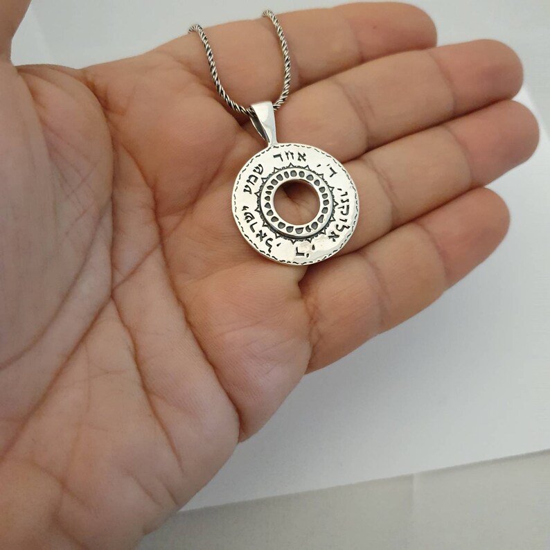 May include: A circular silver pendant featuring a central cutout and Hebrew text engraved along the outer rim. The pendant is suspended from a twisted silver chain, showcasing a metallic finish with dark oxidized detailing within the lettering.