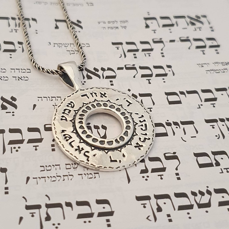 May include: A circular silver pendant on a twisted silver chain. The pendant features a central open circle surrounded by a textured border and an outer ring engraved with Hebrew text. The metal has a polished finish with darkened recessed areas.