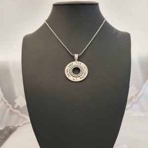 May include: A silver-toned circular pendant featuring a central cutout and Hebrew text engraved along the outer rim. The pendant hangs from a matching silver-toned chain, displaying a textured metallic finish with dark oxidation in the lettering.