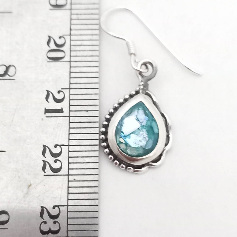 May include: A single silver earring with a teardrop-shaped, light blue glass pendant. The pendant is framed by a beaded silver border and a scalloped edge. The earring hook is also silver.