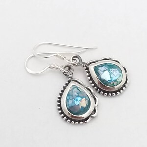 May include: A pair of silver teardrop earrings with a light blue, mosaic-like center. The earrings have a beaded border and a simple hook closure. The light blue color is a prominent feature of the earrings.