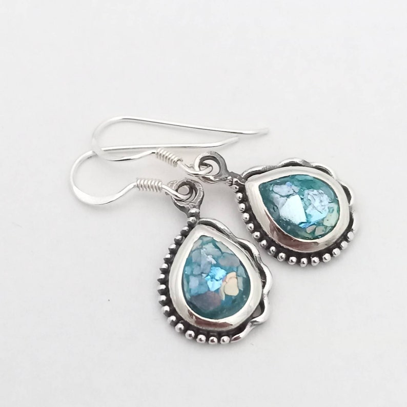 May include: A pair of silver teardrop earrings with a light blue, mosaic-like center. The earrings have a beaded border and a simple hook closure. The light blue color is a prominent feature of the earrings.