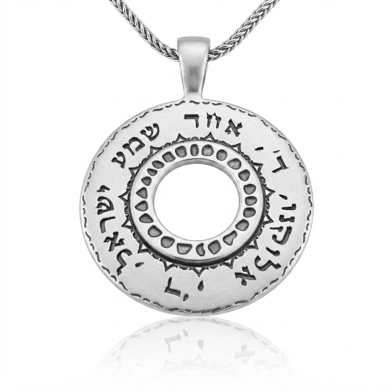 May include: A circular silver pendant featuring Hebrew text engraved along the outer rim and a decorative inner ring with small circular indentations. The pendant is suspended from a silver chain, showcasing a brushed metallic finish throughout.