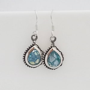 May include: A pair of silver teardrop earrings. Each earring features a bezel-set, aqua-colored glass stone with a mosaic appearance. The silver setting has a beaded border and a decorative loop for the ear wire.