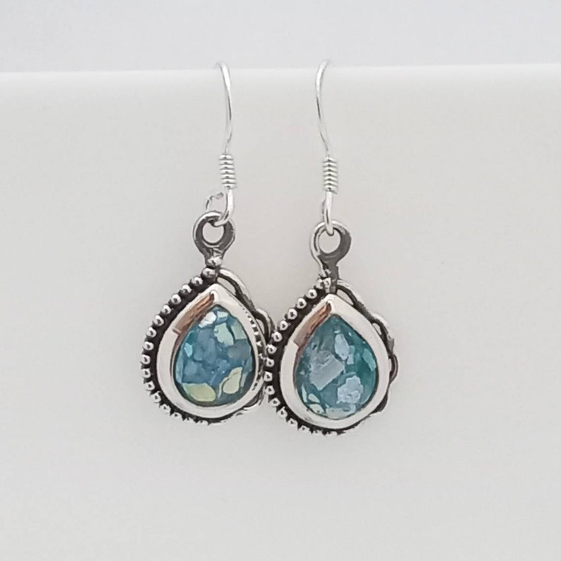 May include: A pair of silver teardrop earrings. Each earring features a bezel-set, aqua-colored glass stone with a mosaic appearance. The silver setting has a beaded border and a decorative loop for the ear wire.