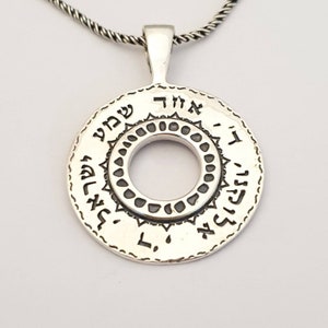 May include: A circular silver pendant featuring Hebrew text engraved along the outer edge and a decorative inner ring with small circular indentations. The pendant is suspended from a twisted silver chain, showcasing a polished metallic finish.