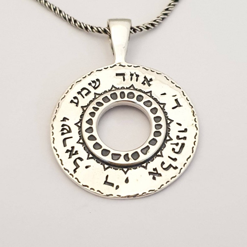 May include: A circular silver pendant featuring Hebrew text engraved along the outer edge and a decorative inner ring with small circular indentations. The pendant is suspended from a twisted silver chain, showcasing a polished metallic finish.