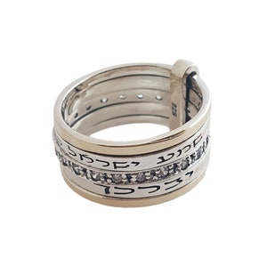 May include: A silver and gold ring with Hebrew text and small clear stones. The ring has multiple bands and a hinged design. The metal is stamped with "925".