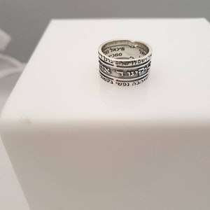 May include: A silver ring with Hebrew text inscribed around the band. The text is in black and reads "ברוך אתה ה' אלוהינו מלך העולם" which translates to "Blessed are you, Lord our God, King of the Universe".