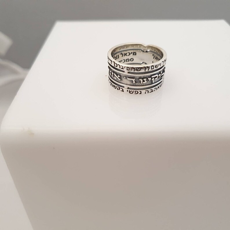 May include: A silver ring with Hebrew text inscribed around the band. The text is in black and reads "ברוך אתה ה' אלוהינו מלך העולם" which translates to "Blessed are you, Lord our God, King of the Universe".