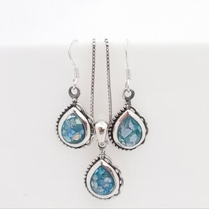 May include: A silver jewelry set featuring a teardrop pendant necklace and matching earrings. Each piece has a silver frame with a beaded edge, surrounding a mosaic of blue and white glass. The set is displayed against a white background.
