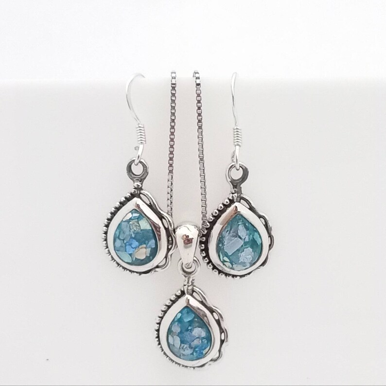 May include: A silver jewelry set featuring a teardrop pendant necklace and matching earrings. Each piece has a silver frame with a beaded edge, surrounding a mosaic of blue and white glass. The set is displayed against a white background.