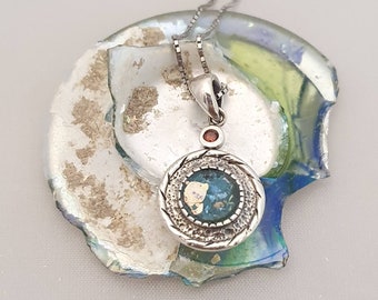 May include: A silver necklace with a round pendant featuring a blue and green Roman glass piece surrounded by a silver frame. The pendant also has a small red gemstone.