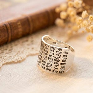 May include: A silver ring with engraved Hebrew text. The ring has a wide band and a polished finish. The text is in black and wraps around the ring's exterior. The ring is marked with "925" on the inside.