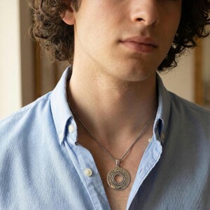 May include: A silver-toned circular pendant necklace featuring engraved Hebrew text around the outer rim and a smaller concentric circle design in the center. The pendant hangs from a thin, twisted silver-toned metal chain against a light blue shirt.