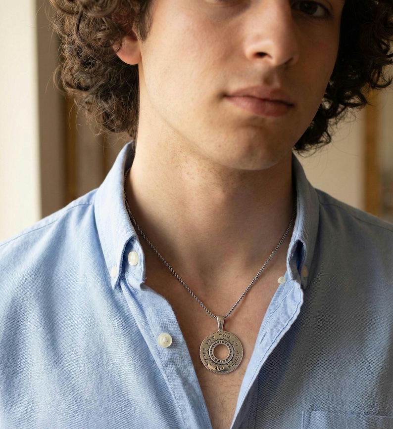 May include: A silver-toned circular pendant necklace featuring engraved Hebrew text around the outer rim and a smaller concentric circle design in the center. The pendant hangs from a thin, twisted silver-toned metal chain against a light blue shirt.