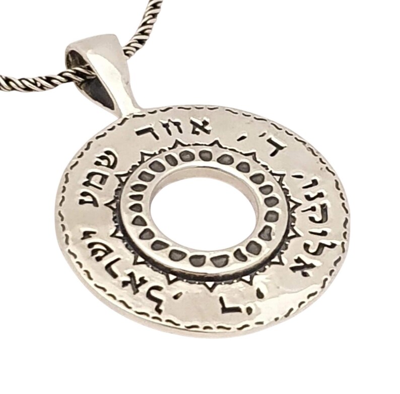 May include: A circular silver pendant on a twisted chain featuring Hebrew text engraved along the outer edge and a decorative border of small indentations surrounding the central circular cutout, creating a polished metallic finish.