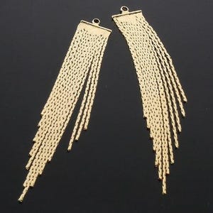 Puede incluir: Dos colgantes de flecos de metal en tono dorado, cada uno con una barra superior rectangular plana con una pequeña anilla circular. Múltiples cadenas finas y texturizadas de diferentes longitudes cuelgan de la barra, creando un efecto en cascada.
