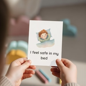 Puede incluir: Una tarjeta con el texto "Me siento seguro en mi cama" sostenida por una persona. La tarjeta presenta una ilustración en acuarela de un niño en la cama. El niño tiene el pelo rojo y está acurrucado en una almohada y una manta azules.