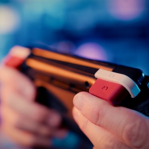 May include: A black handheld gaming controller featuring a prominent red L2 trigger button and a white accent piece. The device has a sleek, dark frame with ergonomic contours designed for grip, highlighting the specific L2 label on the red button.