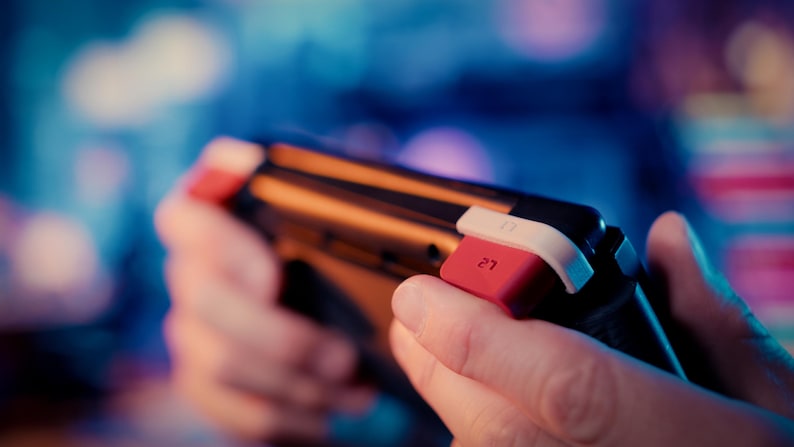 May include: A black handheld gaming controller featuring a prominent red L2 trigger button and a white accent piece. The device has a sleek, dark frame with ergonomic contours designed for grip, highlighting the specific L2 label on the red button.