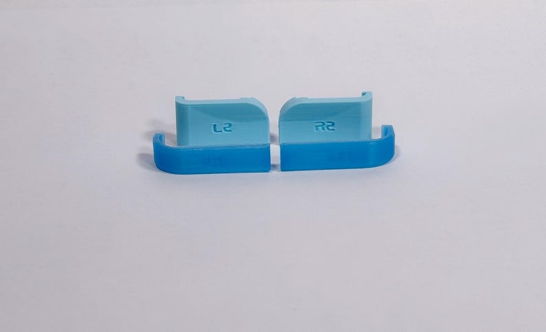 Puede incluir: Dos extensiones de gatillo para mando en color azul claro con acabado liso. La pieza izquierda tiene grabado el texto L2 y la derecha el texto R2. Estos componentes presentan un dise&ntilde;o curvo con una secci&oacute;n base en color azul oscuro.