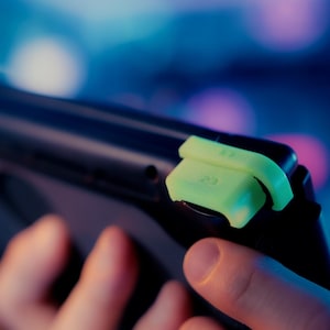 May include: A black handheld gaming console featuring bright neon yellow shoulder buttons. The top edge of the device displays the labels L2 and R2 clearly embossed on the neon yellow plastic triggers, which are attached to the console's frame.
