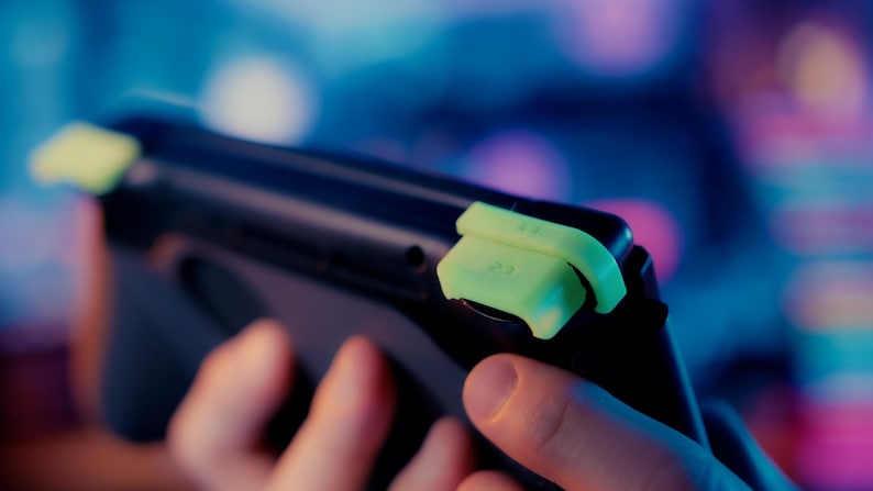 May include: A black handheld gaming console featuring bright neon yellow shoulder buttons. The top edge of the device displays the labels L2 and R2 clearly embossed on the neon yellow plastic triggers, which are attached to the console's frame.