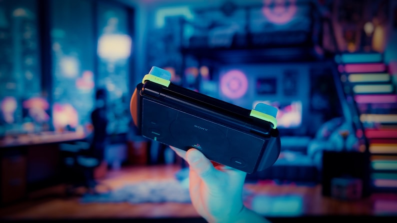 May include: A black handheld gaming console featuring a rectangular body with a Sony logo, integrated side grips, and prominent light blue and lime green shoulder buttons positioned along the top edge of the device for tactile control.