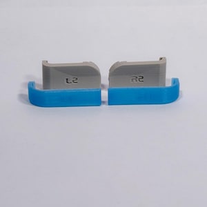 Puede incluir: Dos extensiones de gatillo para mando, una gris y otra azul, con el texto L2 y R2 respectivamente. Estos componentes tienen una forma curva con un borde superior plano y muestran una textura en capas t&iacute;pica de las piezas de pl&aacute;stico impresas en 3D.